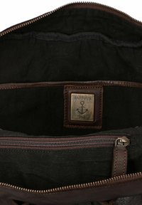 Interior de bolso de cuero marrón con forro de tela negra, cuenta con una etiqueta con un logo de ancla y un compartimento con cremallera. Costura texturizada visible.