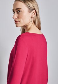 Maglietta aderente a maniche lunghe in tessuto fucsia brillante, con scollatura tonda. Modella mostrata di profilo, con capelli lunghi e orecchini a cerchio.