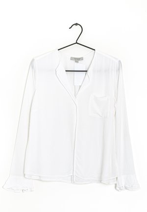 Weiße Langarmbluse mit kleinem Fronttasche an einem schwarzen Kleiderbügel, mit V-Ausschnitt und Rüschenmanschetten.