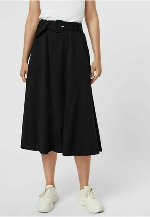 Kobieta w czarnej, midi spódnicy z wysokim stanem i paskiem oraz białych sneakersach, stojąca na jednolitym tle.