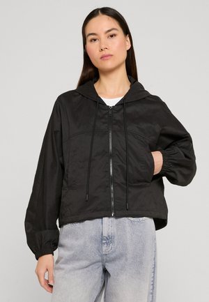 Vrouw met een zwart capuchonjack met rits en elastische manchetten, en lichtblauwe spijkerbroek, staand met één hand in de jaszak.