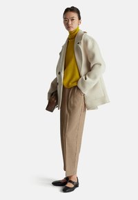 Cappotto di lana crema, dolcevita giallo vivace, pantaloni a quadri marroni e scarpe nere con cinturini. Include una piccola borsa marrone.