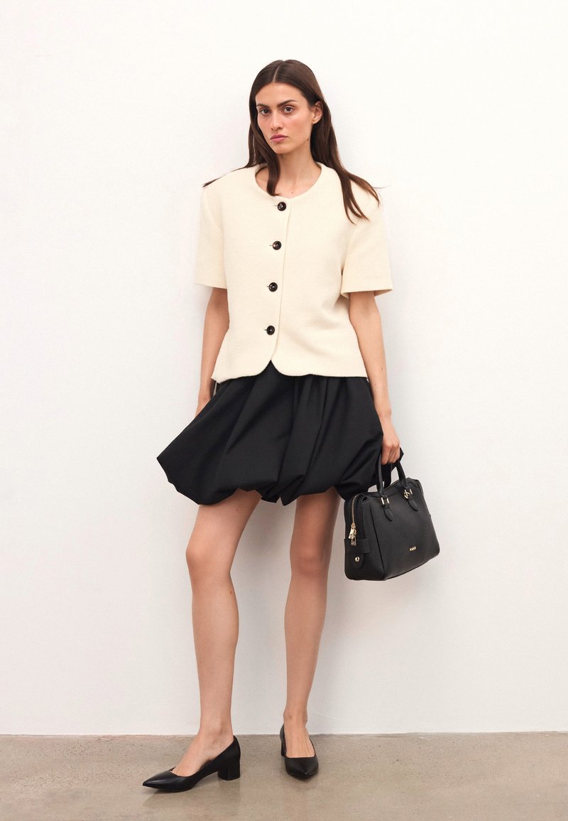 Kreemivärvi lühikeste varrukatega jakk mustade nööpidega, kombineerituna musta volüümika seeliku ja musta käekotiga. Mustad teravatooled kontsad.