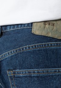 Jeans in denim di un blu scuro, con una patch di pelle visibile etichettata "EDWIN". Cuciture di colore oro a contrasto lungo le cuciture.