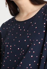 Blusa di un blu scuro realizzata in morbido tessuto, con un motivo geometrico rosso e bianco sparso. Collo rotondo e maniche rilassate.