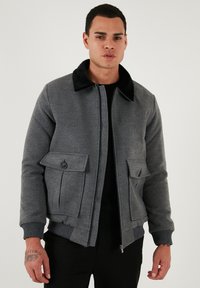 Sivý bombardér z textúrovaného materiálu s čiernym kožúškom na golieri, dvoma klopovými vreckami a rebrovanými manžetami a lemom. Zapínanie na zips vpredu.