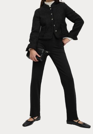 Femme portant une veste noire boutonnée, un pantalon noir et des ballerines noires, tenant une pochette noire d'une main sur la hanche.