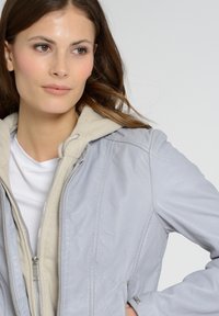 Lichtgrijze leren jas met een rits aan de voorkant, gewatteerd ontwerp en een beige capuchon. Heeft subtiele stiksels en zilveren hardware accenten.