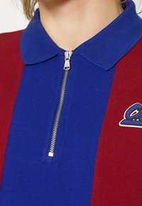 Gros plan sur le col d'une chemise zippée à rayures verticales bleues et bordeaux avec un logo partiel sur le côté droit.