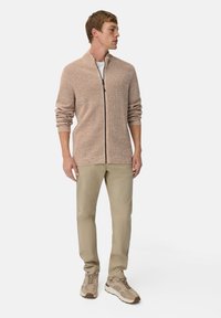 Strickjacke in hellbeige mit Reißverschluss, umgeschlagenem Kragen und gerippten Bündchen. Kombiniert mit beigen Hosen und strukturierten Sneakern.