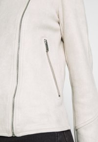 Jasnobeżowa kurtka z imitacji zamszu z ukośnym zamkiem błyskawicznym, boczną kieszenią zamykaną na zamek oraz gładką fakturą.