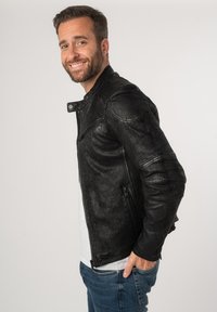 Zwarte leren jack met een getextureerde afwerking, slim fit, met rits aan de voorkant en schouderdetails. Gekleed met een wit shirt en blauwe jeans.