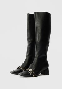 Black leather knee-high boots with a block heel, featuring a pointed toe and a buckle strap adorned with studs on the ankle.