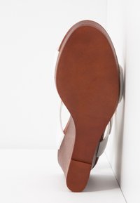 Semelle marron d'une sandale blanche à talon carré, posée sur une surface blanche réfléchissante sur fond blanc.