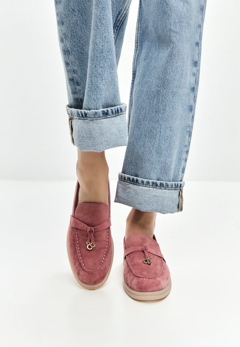 Pieds portant des mocassins en daim rose avec des détails dorés, associés à un jean en denim bleu clair retroussé sur fond blanc.