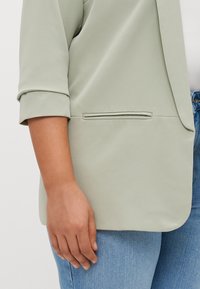 Lichtgroene blazer met een gladde textuur, ontspannen pasvorm, opgerolde mouwen en een zijzak, gecombineerd met lichtblauwe jeans.