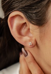 Gold flower-shaped stud earring with three small sparkling gems arranged in petals, set against a close-up of the ear.
