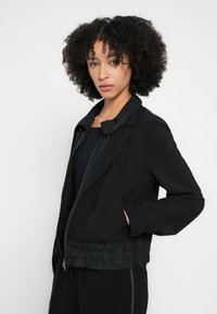 Jachetă bomber neagră cu talie și manșete ribbed, având un fermoar frontal și material ușor texturat. Design simplu al gulerului, fără ornamente.