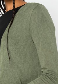 Cárdigan verde oliva de punto ligero y texturizado. Presenta un diseño de frente abierto y mangas largas, superpuesto a una blusa negra.