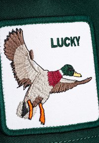 Zelena kapica s pravokutnim bijelim panelom na kojem je izvezeni patak u letu, obojan smeškom, sivom i burgundskom, s riječju "LUCKY" iznad.