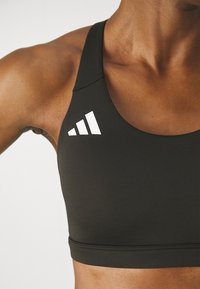Svart sports-bh med en jevn tekstur og racerback-design. Har hvit logo med tre striper på venstre skulder.