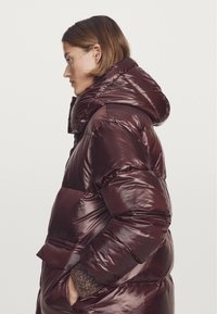 Vrouw in profiel die een glanzende donker-bordeaux kleurige pufferjack met een grote capuchon en gebreide mouwen draagt, met haar handen in de zakken van de jas.