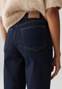 Dark blue denim jeans with a textured finish, featuring a tan leather brand patch and yellow stitching detail on the back pockets.