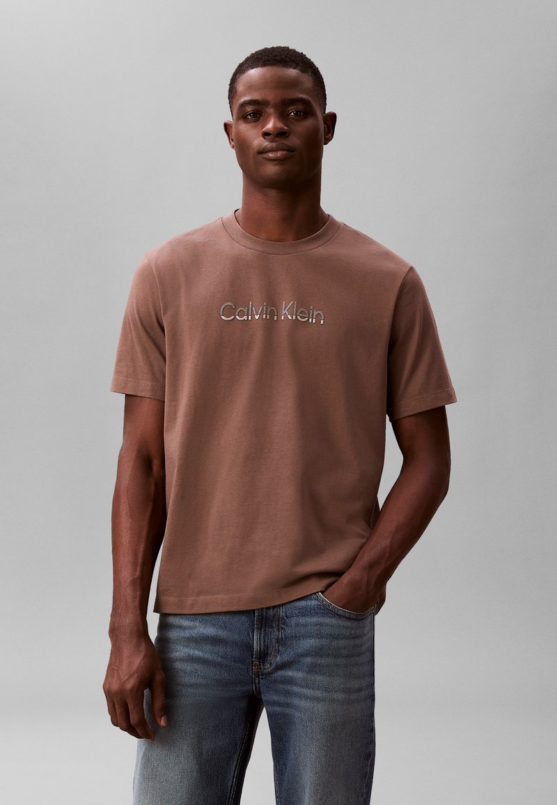 Άνδρας φορώντας καφέ μπλούζα Calvin Klein και μπλε τζιν, στέκεται με το ένα χέρι στην τσέπη μπροστά σε απλό γκρι φόντο.