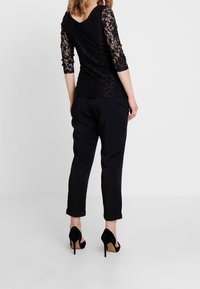 Schwarzes Spitzenshirt mit Dreiviertelärmeln und V-Ausschnitt, kombiniert mit schwarzen, umgeschlagenen Hosen und schwarzen High-Heels.