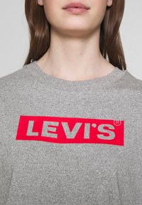 Camiseta de algodón gris con un destacado logo rectangular rojo que lleva el texto "LEVI'S®" en blanco, con escote redondo y mangas cortas.