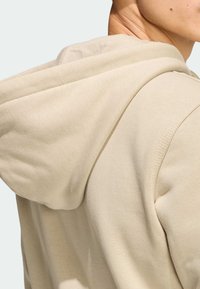 Béžová mikina s kapucňou ukazujúca textúru a švy, s jemnou tkaninou padajúcou cez rameno. Viditeľná kapucňa a čisté švy.