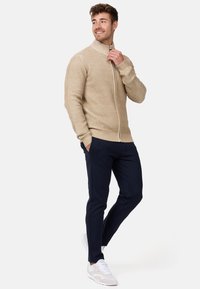 Sudadera beige con cremallera y punto texturizado, combinada con pantalones slim-fit azul marino y sneakers claros. La sudadera cuenta con puños y cuello de canalé.