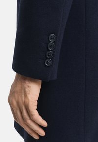 Primo piano di una mano accanto alla manica di un cappotto blu scuro con quattro bottoni neri disposti in fila verticale vicino al polsino.