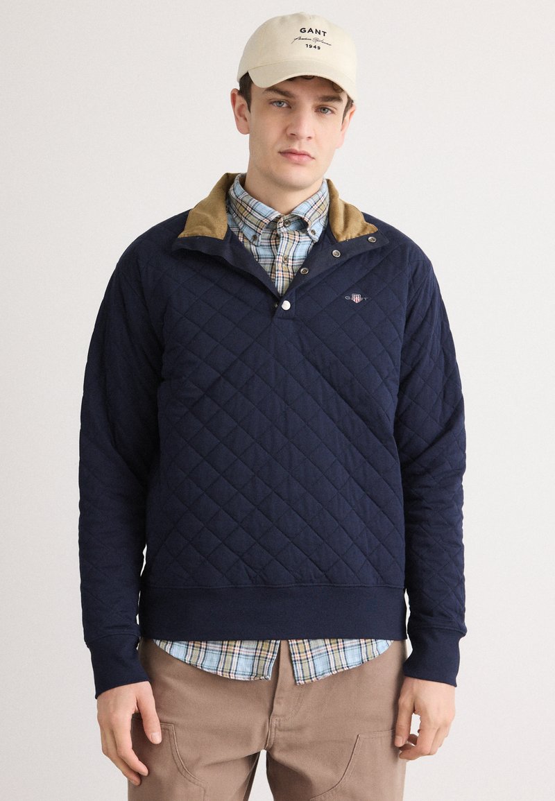 Marineblauwe gewatteerde pullover met een tan kleurige kraag, voorzien van een half-knopenlijst en een logo detail. Draagt over een blauwe en geruite overhemd met knopen.