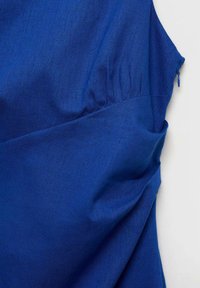 Haut en tissu léger bleu avec une coupe décontractée, des plis latéraux et un éclat subtil. Comprend une fermeture éclair dissimulée à l'épaule.