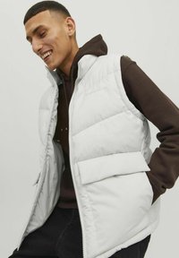 White padded vest with a front zipper and large pocket, worn over a dark brown hoodie. The vest features a matte fabric and quilted design.