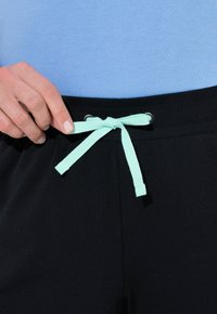 Czarne spodnie z prążkowanym pasem, wyposażone w jasnoszmaragdowy sznurek ściągający. Ręka trzyma sznurek na tle niebieskiego topu.