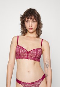 Burgundy spetsbalkong med genomskinlig blommönster, vågiga kanter och justerbara band, med en liten rosett i mitten fram.