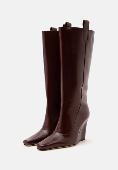 Paire de bottes compensées hautes en cuir marron foncé avec bouts pointus et languettes en haut, sur un fond clair uni.
