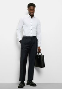 Weißes Langarmhemd mit schwarzen Knöpfen, kombiniert mit navyblauen Anzughosen. Schwarze Schuhe und eine schwarze Aktentasche runden das Outfit ab.