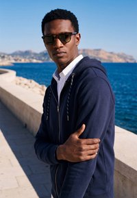 Homme portant des lunettes de soleil et un sweat à capuche bleu marine debout près d'une promenade en bord de mer avec des montagnes et de l'eau bleue en arrière-plan lors d'une journée ensoleillée.
