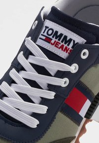 Sneaker in tela blu navy con lacci bianchi, caratterizzato da un pannello verde oliva testurizzato e una toppa con logo a strisce rosse, bianche e blu.