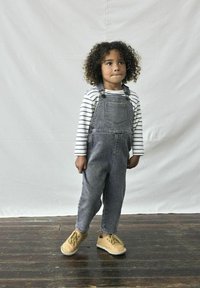 Salopette en jean grise avec des bretelles réglables, accompagnée d'un T-shirt à manches longues rayé blanc et bleu marine et de chaussures tan à lacets.