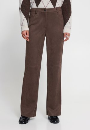 Pantalon marron à jambes droites porté avec un pull à losanges blanc et marron et des chaussures en cuir noires, mains reposant le long du corps avec des bagues aux doigts.