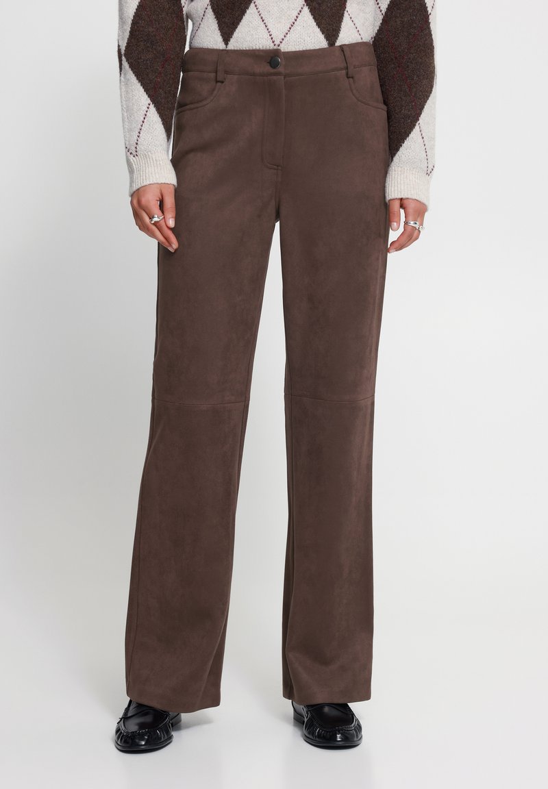 Pantalon marron à jambes droites porté avec un pull à losanges blanc et marron et des chaussures en cuir noires, mains reposant le long du corps avec des bagues aux doigts.