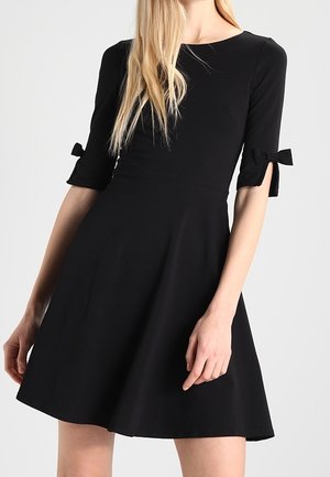 Femme portant une robe noire jusqu'aux genoux avec des manches aux coudes, chacune présentant un petit détail nœud près de l'ourlet.