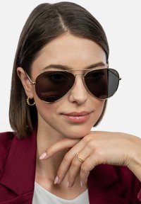 Pilotní sluneční brýle s tmavými čočkami, zlatým kovovým rámem a jemnými detaily. Modelka má neutrální výraz a na sobě má vínový blejzr.
