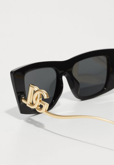 Black acetate sunglasses with gray lenses, featuring a gold metal chain and logo accent attached to the frame.