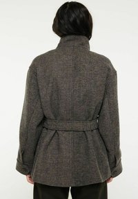 Grå kappa med fiskbensmönster, hög krage, bälte i midjan och knäppta manschetter, tillverkad av strukturerad ullblandning.