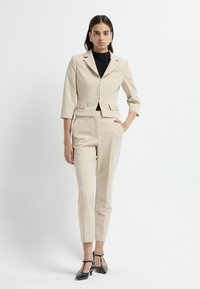 Béžový zkrácený blazer s vypasovaným designem a dvěma předními kapsami, spárovaný s odpovídajícími tapered kalhotami a černými lodičkami na vysokém podpatku.